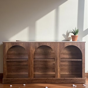 May include: A wooden cabinet with three arched openings, each with a shelf. The cabinet is made of dark brown wood and has a light-colored top. A small potted plant sits on top of the cabinet. The cabinet is on a wooden floor.