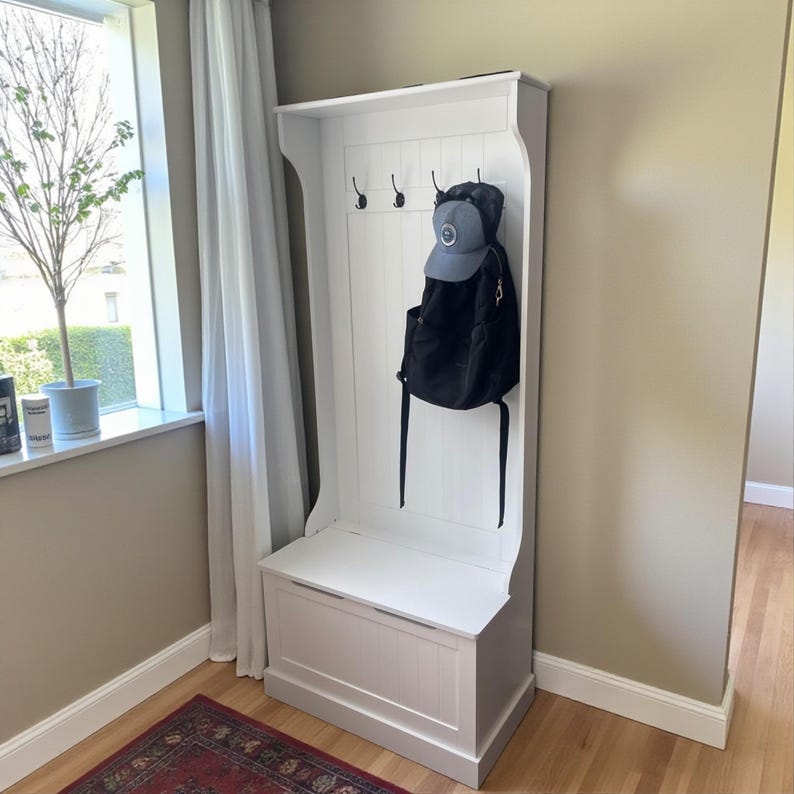 Puede incluir: Un mueble recibidor de madera blanca con un banco y un compartimento de almacenamiento. Cuenta con un tablero trasero con ganchos para colgar objetos y un estante en la parte superior. Una mochila negra y una gorra de b&eacute;isbol gris cuelgan de los ganchos.