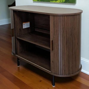 May include: A mid-century modern style cabinet with a light wood top and dark wood body. The cabinet has a curved front with vertical wood slats and a sliding door. It features a shelf inside and sits on black legs.