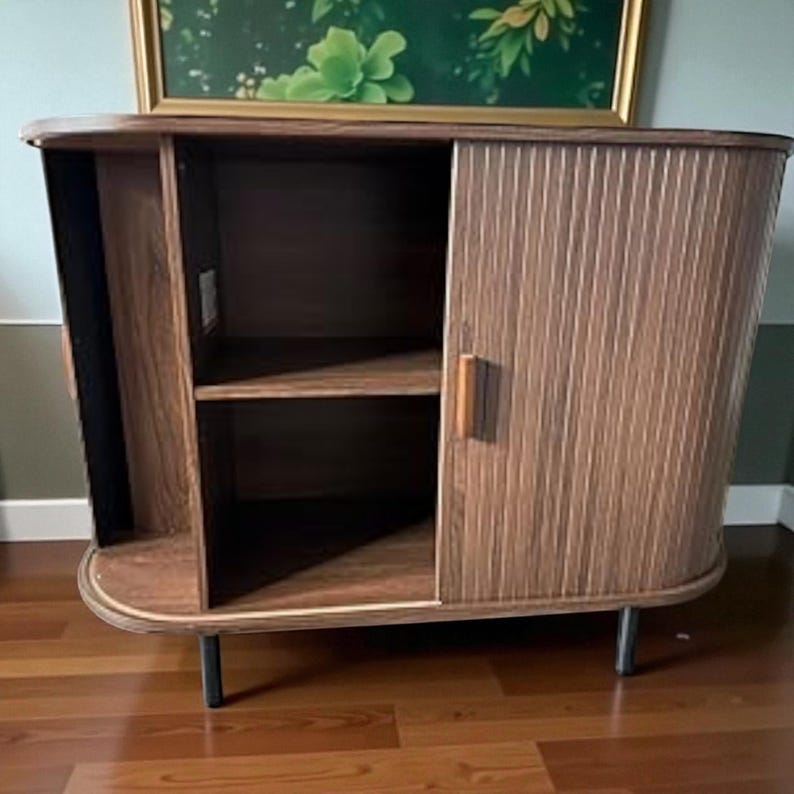 May include: A mid-century modern style cabinet with a curved design. The cabinet is made of wood with a vertical striped pattern on the sliding door. It has a shelf inside and sits on black metal legs.