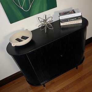 May include: A black wooden cabinet with a curved design, featuring a decorative gold metal flower and a stack of books. A cream-colored bowl with two black objects sits on the cabinet's surface. The cabinet is set against a white wall with a green painting.