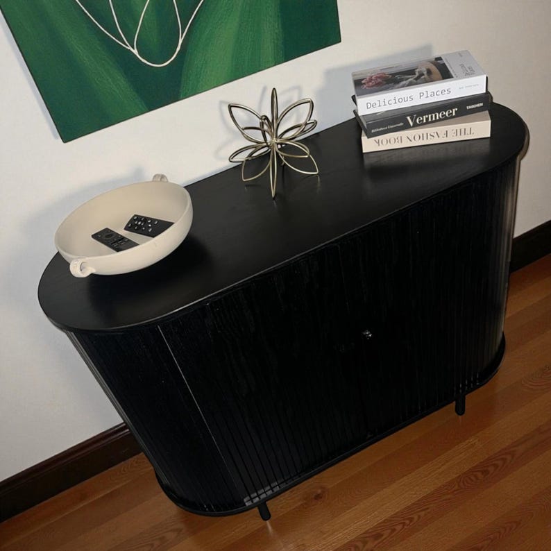 May include: A black wooden cabinet with a curved design, featuring a decorative gold metal flower and a stack of books. A cream-colored bowl with two black objects sits on the cabinet's surface. The cabinet is set against a white wall with a green painting.