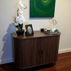May include: A dark brown wooden cabinet with a curved top and vertical grooved detailing on the doors. The cabinet is adorned with a black framed photo, a decorative tray, and a white orchid in a black pot. A green abstract painting hangs above.