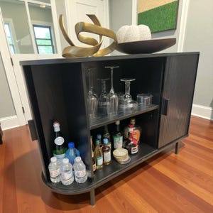 May include: A black bar cabinet with a curved front and two sliding doors. The cabinet has two shelves, one with wine glasses and the other with bottles of liquor. The top of the cabinet has a gold sculpture and a decorative bowl.