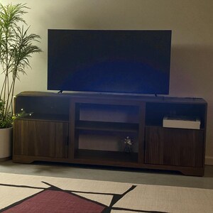 May include: A dark brown wooden TV stand with two cabinet doors, open shelving, and a large flat-screen TV on top. A potted plant is visible on the left side. The stand has a modern design and is placed on a patterned rug.