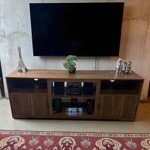 May include: A dark brown wooden TV stand with a large black television mounted above it. The stand has two doors with vertical wood grain, open shelves, and a digital clock displaying 8:50. Decorative items include a small Eiffel Tower and a bonsai tree.