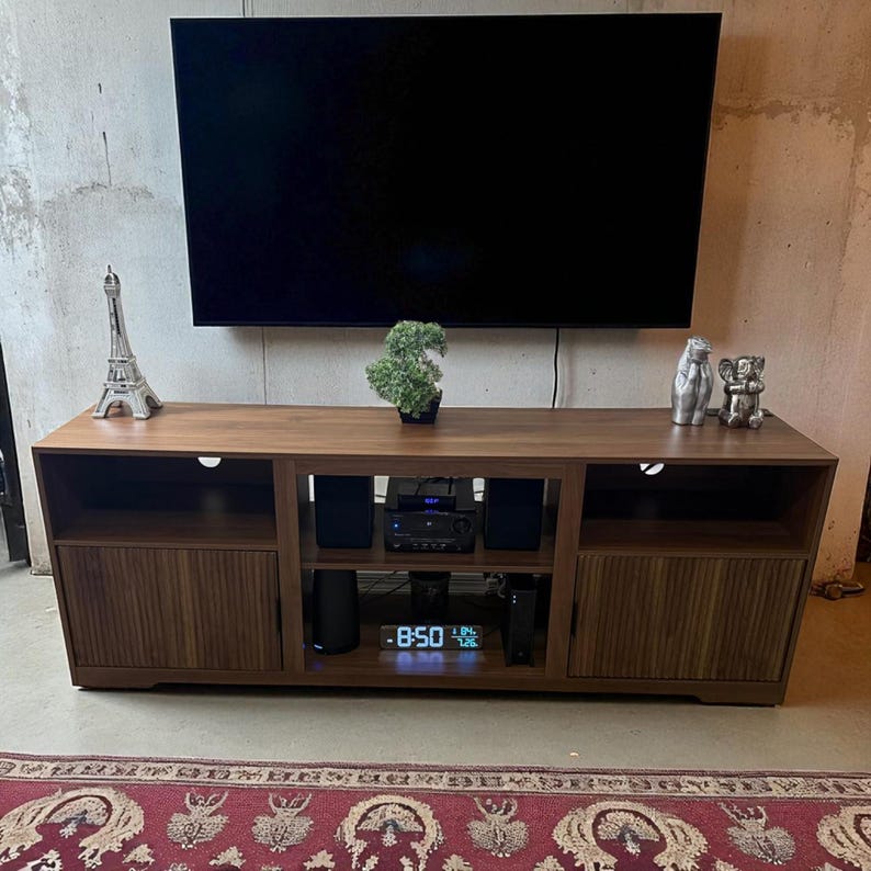 May include: A dark brown wooden TV stand with a large black television mounted above it. The stand has two doors with vertical wood grain, open shelves, and a digital clock displaying 8:50. Decorative items include a small Eiffel Tower and a bonsai tree.