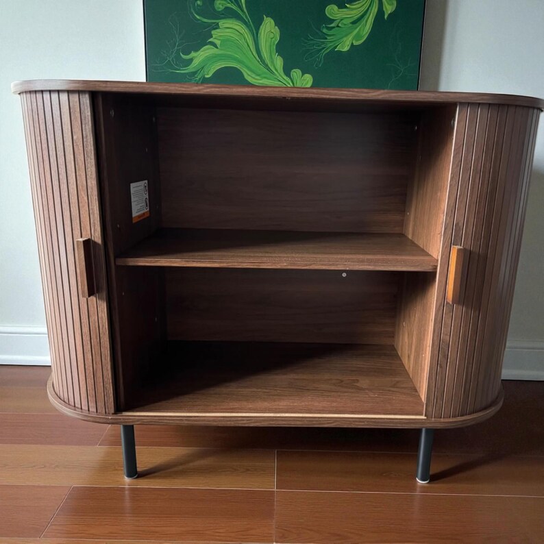May include: A dark brown wooden cabinet with a curved front and vertical grooved detailing. The cabinet has two doors with wooden handles, a shelf, and black metal legs. The interior is a lighter shade of brown.