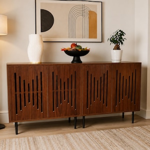 May include: A mid-century modern style wooden cabinet with a dark brown finish and black legs. The cabinet doors feature vertical cut-out designs. A white vase, a black bowl with decorative items, and a potted plant sit on top. A framed abstract art piece hangs on the wall.