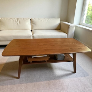 May include: A rectangular wooden coffee table with a natural wood grain finish. The table has a lower shelf and angled legs. The table is approximately 48 inches long and 24 inches wide.