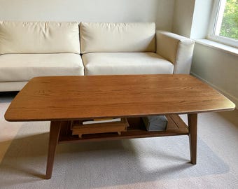 May include: A rectangular wooden coffee table with a natural wood grain finish. The table has a lower shelf and angled legs. The table is approximately 48 inches long and 24 inches wide.