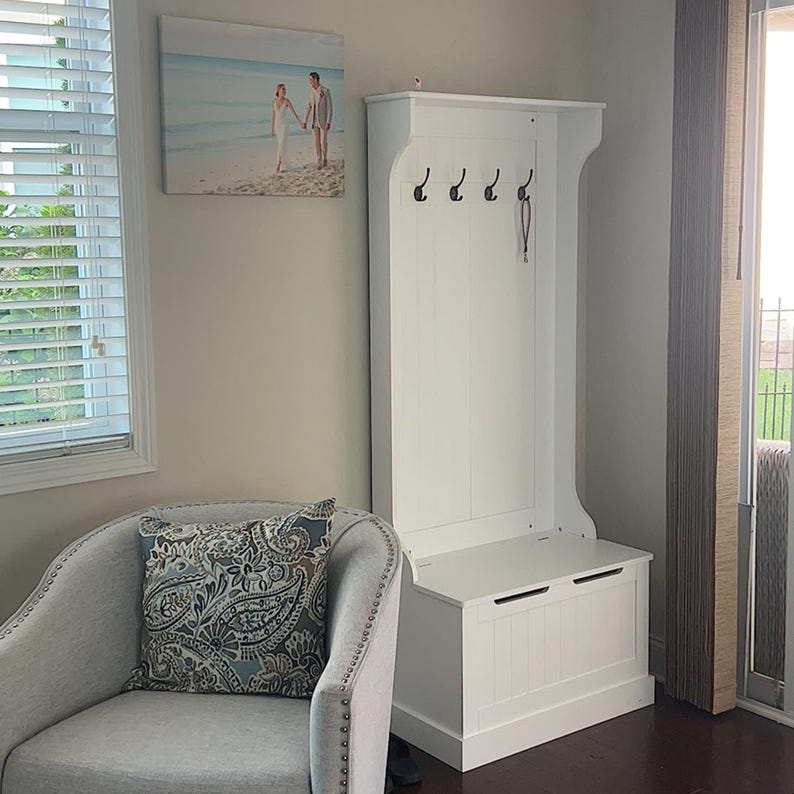 Puede incluir: Un mueble recibidor de madera blanca con asiento y ganchos para abrigos. Una foto enmarcada de una pareja en la playa cuelga de la pared. Un sill&oacute;n gris con un coj&iacute;n estampado est&aacute; en primer plano.
