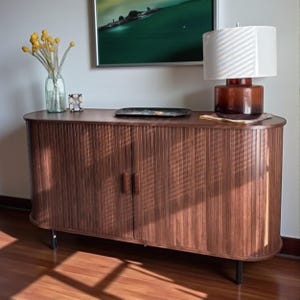May include: A wooden cabinet with a ribbed front and two doors. The cabinet has a curved top and sits on black legs. A lamp with a white shade and a brown base is on top, along with a decorative tray and a vase of yellow flowers.