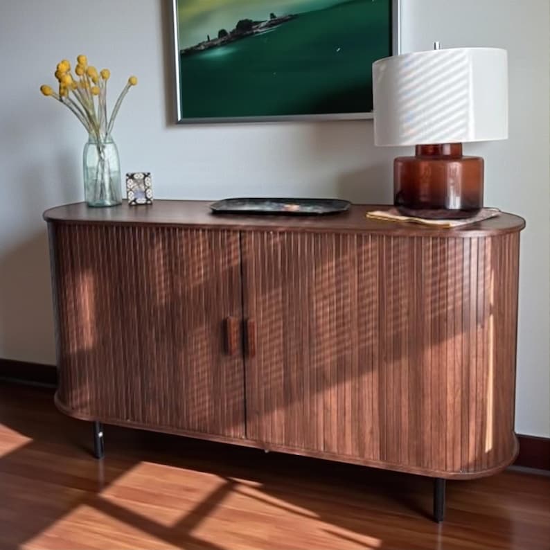 May include: A wooden cabinet with a ribbed front and two doors. The cabinet has a curved top and sits on black legs. A lamp with a white shade and a brown base is on top, along with a decorative tray and a vase of yellow flowers.