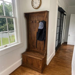 Puede incluir: Un banco de entrada y perchero de madera de color marr&oacute;n oscuro. El banco tiene un compartimento de almacenamiento. El perchero tiene varios ganchos y una balda. Una mochila negra y una gorra de b&eacute;isbol azul cuelgan de los ganchos. Un reloj est&aacute; colgado en la pared.
