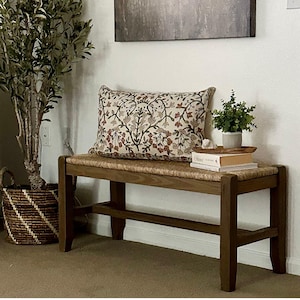 May include: A wooden bench with a woven seat and dark brown legs. A decorative pillow with a floral pattern rests on the bench. A small potted plant and books are placed on the bench, with a large plant in a basket to the left.