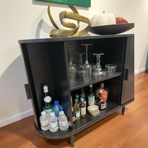 May include: A black bar cabinet with a curved front and legs. The cabinet has two shelves, displaying glassware, bottles of liquor, and bottled water. A gold sculpture and a decorative bowl with a white pumpkin and red fruit sit on top.
