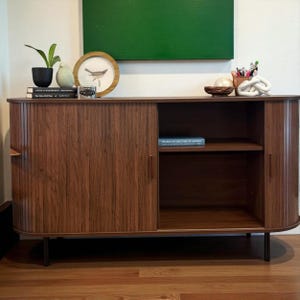May include: A mid-century modern style wooden cabinet with sliding doors. The cabinet has a dark brown finish, black legs, and a shelf inside. Decorative items on top include a framed bird print, books, and a small plant.
