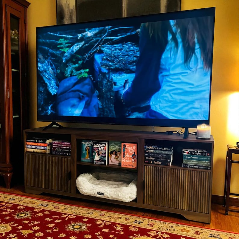 May include: A dark brown wooden TV stand with a large flat-screen television on top. The stand has multiple shelves filled with books, a pet bed, and a lit candle. A red patterned rug is in the foreground.