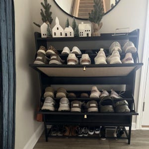 May include: A black shoe rack with multiple shelves, filled with various shoes. The rack is against a white wall, with decorative miniature houses and trees on top. A round mirror is visible in the background.