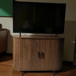 May include: A dark, flat-screen television sits atop a wooden cabinet. The cabinet has a ribbed design on the doors and is supported by black metal legs. The cabinet is a warm brown color, and the television screen is black.