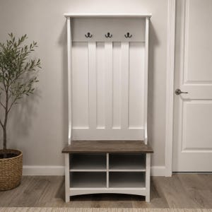 May include: A white hall tree featuring a dark wood-toned bench seat, a vertical back panel with three black double-pronged metal hooks, and a lower storage base divided into four open cubby compartments for organized storage of footwear or items.