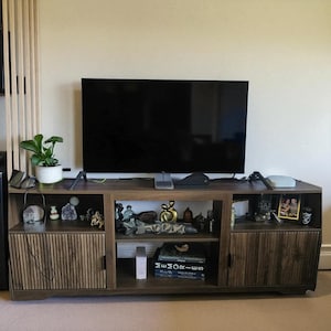 May include: A dark brown wooden TV stand with a large black screen television on top. The stand has two doors with vertical grooves and open shelves displaying decorative items, books, and a small potted plant.