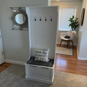 Puede incluir: Un banco de entrada de madera blanca con un compartimento de almacenamiento, un asiento acolchado y cuatro ganchos negros. Un coj&iacute;n decorativo con el texto "LIVE SIMPLY, DREAM BIG, BE GRATEFUL, GIVE LOVE, LAUGH LOTS" descansa sobre el asiento.