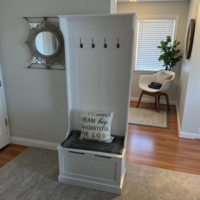 Puede incluir: Un banco de entrada de madera blanca con un compartimento de almacenamiento, un asiento acolchado y cuatro ganchos negros. Un coj&iacute;n decorativo con el texto "LIVE SIMPLY, DREAM BIG, BE GRATEFUL, GIVE LOVE, LAUGH LOTS" descansa sobre el asiento.