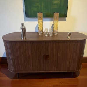 May include: A dark brown wooden cabinet with a curved top and vertical grooved door panels. It holds a cocktail shaker, two bottles, three small glasses, and a small gold-colored cup. The cabinet sits on thin, dark metal legs.