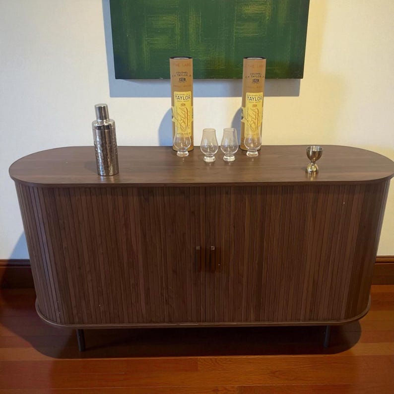 May include: A dark brown wooden cabinet with a curved top and vertical grooved door panels. It holds a cocktail shaker, two bottles, three small glasses, and a small gold-colored cup. The cabinet sits on thin, dark metal legs.