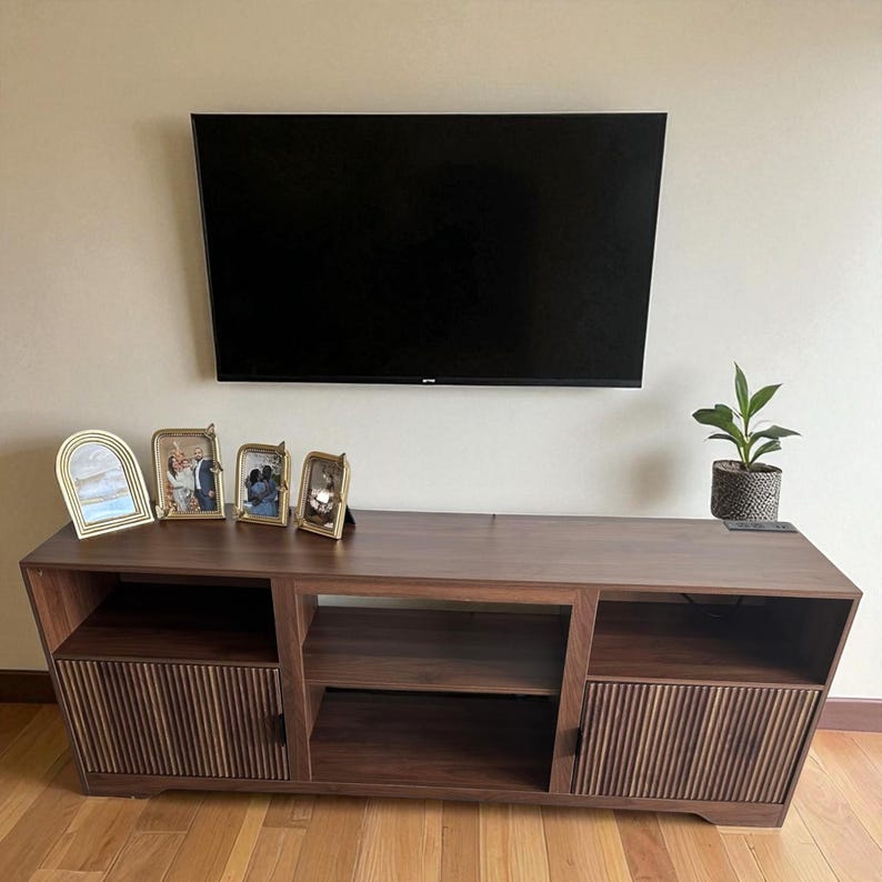 May include: A dark brown wooden TV stand with a black television mounted above it. The stand has two cabinets with vertical wood paneling and an open shelf. Decorative gold picture frames and a potted plant sit on the stand.