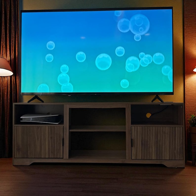 May include: A large flat-screen television displaying a blue and green gradient with white bubbles, set atop a wooden entertainment center. The center has two doors with vertical grooves and open shelving.