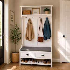 May include: A white entryway bench with a gray cushion, two drawers, and a shoe rack. Above, there's a shelf with a woven basket, a sign that says "welcome home", and a potted plant. Coats and a purse hang from hooks.