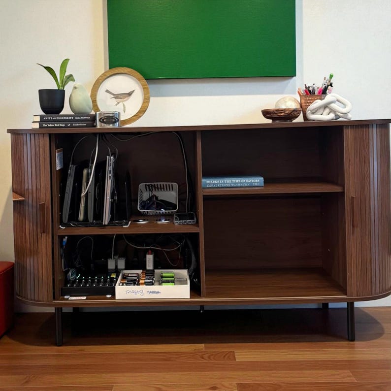 May include: A mid-century modern style wooden cabinet with a dark brown finish. The cabinet has two doors with vertical slat detailing and black legs. The interior has shelves and is filled with electronics, books, and decorative items. A green painting hangs above.