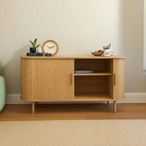 May include: A light oak wood cabinet with a curved design, featuring a sliding door on the left and an open shelf on the right. The cabinet is raised on short, tapered legs. Decorative items, including a clock and a plant, are displayed on top.