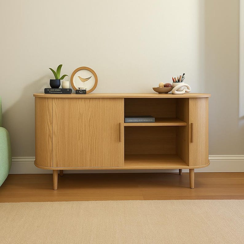 May include: A light oak wood cabinet with a curved design, featuring a sliding door on the left and an open shelf on the right. The cabinet is raised on short, tapered legs. Decorative items, including a clock and a plant, are displayed on top.