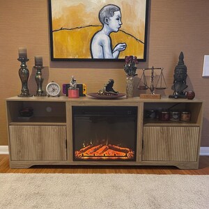 May include: A light wood-tone entertainment center with a built-in electric fireplace. The center features two storage cabinets with vertical grooved doors, open shelving, and a display of decorative items, including candles, a clock, and a Buddha statue.