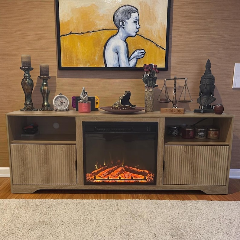 May include: A light wood-tone entertainment center with a built-in electric fireplace. The center features two storage cabinets with vertical grooved doors, open shelving, and a display of decorative items, including candles, a clock, and a Buddha statue.