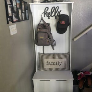 Puede incluir: Un organizador de entrada blanco con un banco, ganchos y un estante. Una mochila gris y una gorra negra cuelgan de los ganchos. Un coj&iacute;n a rayas con la palabra "family" descansa sobre el banco. Un letrero negro de "hello" est&aacute; arriba.