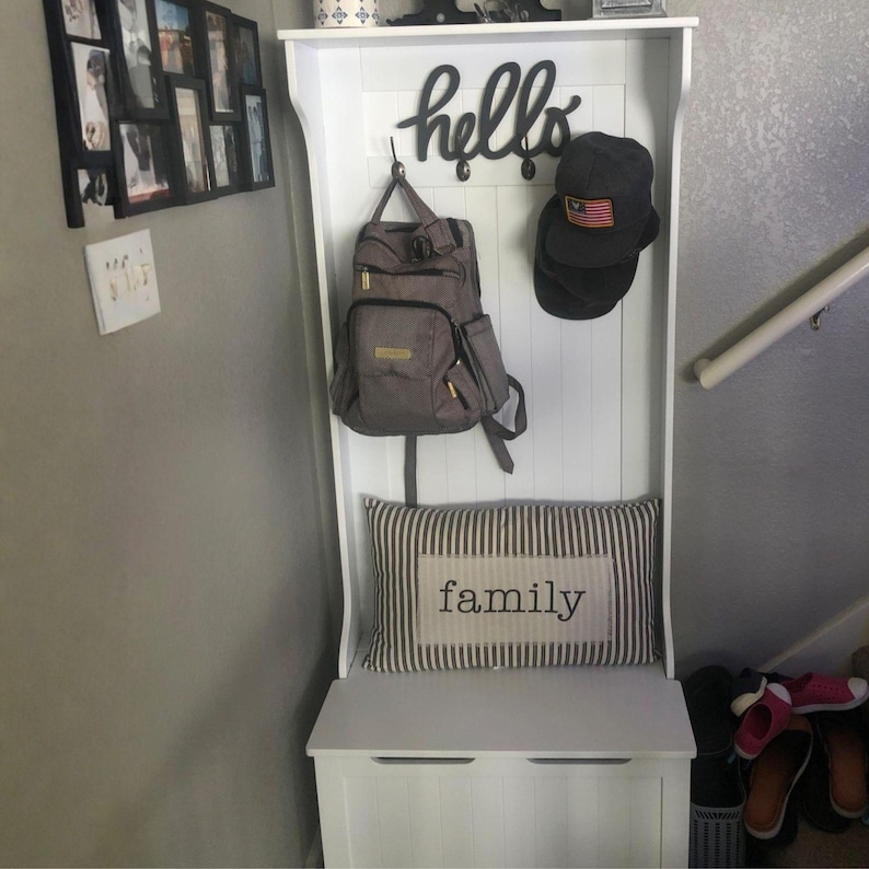 Puede incluir: Un organizador de entrada blanco con un banco, ganchos y un estante. Una mochila gris y una gorra negra cuelgan de los ganchos. Un coj&iacute;n a rayas con la palabra "family" descansa sobre el banco. Un letrero negro de "hello" est&aacute; arriba.
