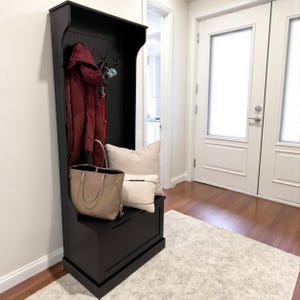 Puede incluir: Un banco de entrada negro con perchero, ganchos y almacenamiento. Un abrigo burdeos cuelga de los ganchos, y un bolso beige y almohadas est&aacute;n sobre el banco. El banco est&aacute; sobre una alfombra de color claro.