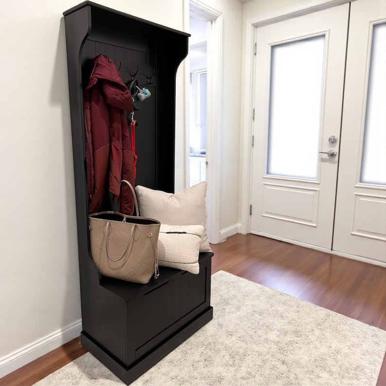 Puede incluir: Un banco de entrada negro con perchero, ganchos y almacenamiento. Un abrigo burdeos cuelga de los ganchos, y un bolso beige y almohadas est&aacute;n sobre el banco. El banco est&aacute; sobre una alfombra de color claro.