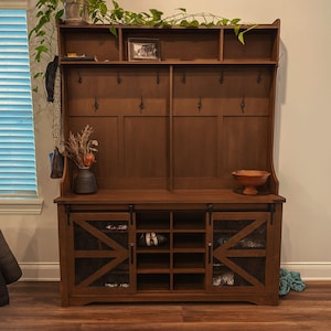 May include: A dark brown wooden entryway organizer with a bench, shoe storage, and coat hooks. The unit has sliding doors with a mesh design and a top shelf for decor. A vase with dried flowers and a wooden bowl are on display.