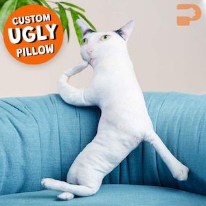 May include: A white cat-shaped pillow with black markings on its head and green eyes, posed thoughtfully on a blue sofa. The pillow has a whimsical design, with the text "CUSTOM UGLY PILLOW" in an orange circle.