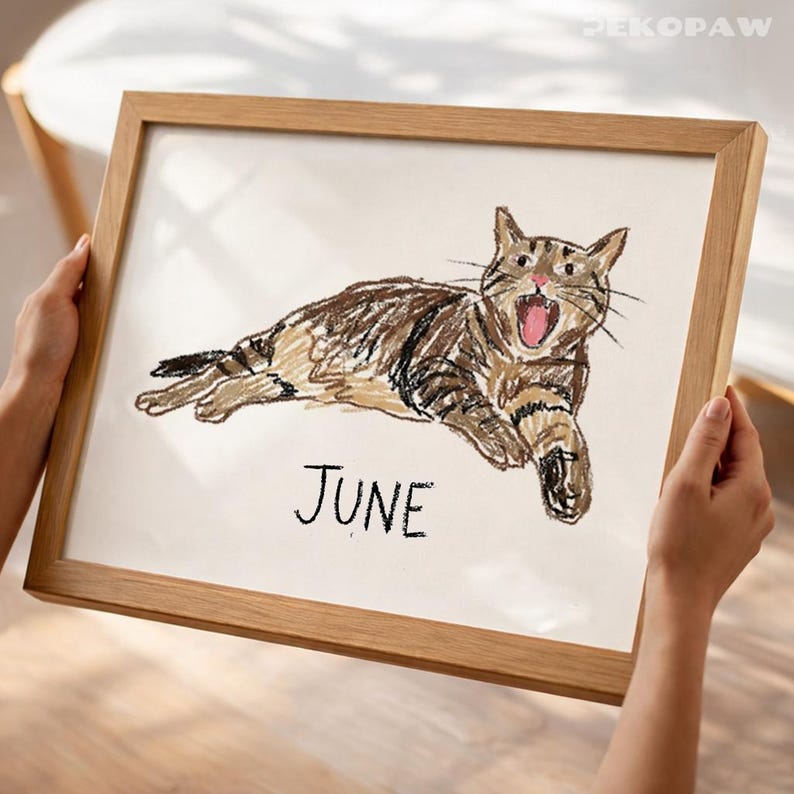 Puede incluir: Una obra de arte enmarcada con una ilustraci&oacute;n de un gato atigrado marr&oacute;n con la boca abierta. La palabra "JUNE" est&aacute; escrita debajo. El marco es de madera clara.