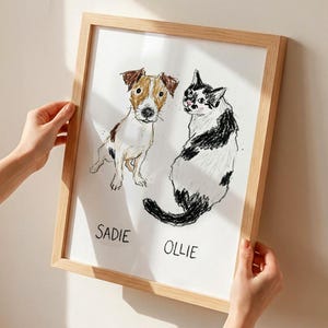 Puede incluir: Una obra de arte enmarcada con bocetos a l&aacute;piz de un perro y un gato. El perro es marr&oacute;n y blanco, y el gato es blanco y negro. Los nombres "Sadie" y "Ollie" est&aacute;n escritos debajo. El marco es de madera clara.