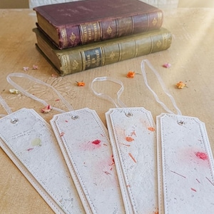 Könnte beinhalten: Vier rechteckige Lesezeichen aus cremefarbenem, strukturiertem Papier mit eingearbeiteten bunten Blumenpartikeln. Jedes Lesezeichen hat einen genähten Rand, eine silberne Metallöse oben und eine dünne, durchscheinende weiße Bandschlaufe.