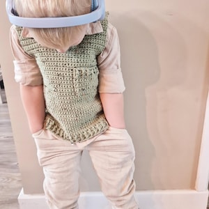 Op de afbeelding: Een kind draagt een lichtgroen gehaakt vest, een overhemd met kraag en een lichtgekleurde broek. Het kind draagt ook een lichtblauwe koptelefoon om de nek en witte en bruine sneakers. Het kind heeft de handen in de zakken.
