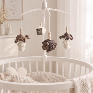 May include: A white crib mobile featuring crocheted hanging ornaments including two brown and white mushrooms and two hedgehogs in shades of brown and cream. The ornaments are suspended by white strings with light wooden beads on a white frame.